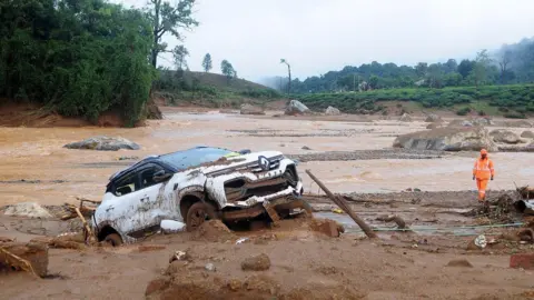 Reuters Seorang penyelamat berjalan melewati mobil yang rusak di lokasi longsor setelah beberapa kali tanah longsor melanda perbukitan di Wayanad.