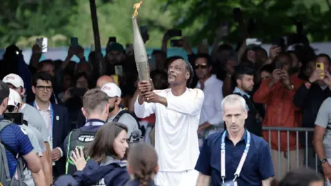 Getty Images Rapper AS Snoop Dogg memegang obor sebagai bagian dari estafet obor Olimpiade Paris 2024 pada upacara pembukaan pada 26 Juli 2024 di Saint-Denis, di luar Paris.