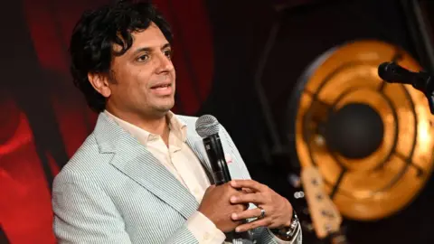 Warner Bros. M. Night Shyamalan mengenakan kemeja putih dan blazer bergaris biru