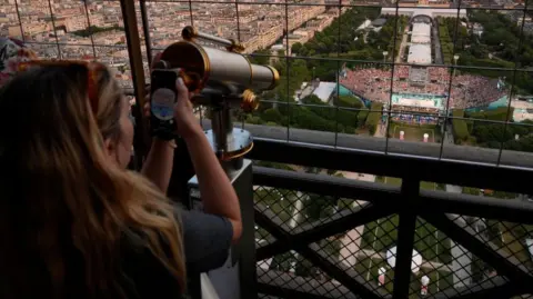 Seorang wanita menggunakan teleskop di Menara Eiffel untuk menonton pertandingan bola voli di stadion di bawah Getty Images