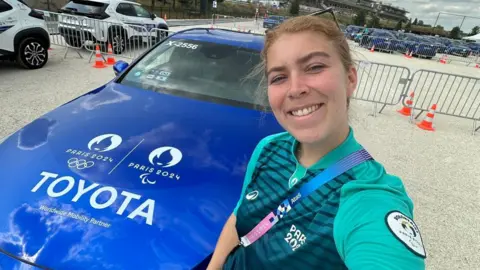 Elisabeth Lomholt berpose selfie di depan mobil sedan berwarna biru tua berlogo Paris 2024 dan branding Toyota. Dia mengenakan seragam sukarelawan resmi. Sekitar dua lusin mobil diparkir di tempat parkir darurat sebagai latar belakang.