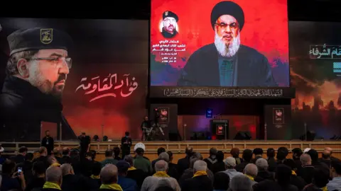 Reuters Pendukung Hizbullah mendengarkan pidato pemimpin kelompok itu Hassan Nasrallah (kanan) pada upacara peringatan komandan senior Fuad Shukr (kiri) setelah dia terbunuh dalam serangan Israel (6 Agustus 2024).