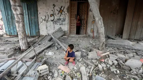 Reuters Seorang anak laki-laki Palestina duduk di lokasi serangan Israel terhadap sebuah rumah di kamp pengungsi Maghazi di Jalur Gaza tengah (14 Agustus 2024).