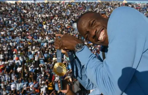     Monirul Bhuyan / AFP Letsile Tebogo berpose dengan medali emas Olimpiade di sebuah stadion di Gaborone, Botswana.