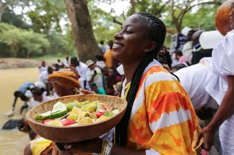 AKINTUNDE AKINLEYE / REUTERS Seorang pemuja Osun memberikan persembahan dan berdoa di labu di tepi Sungai Osun selama festival tahunan Osun di Hutan Suci Osun-Osogbo di Osogbo, barat daya Nigeria, 9 Agustus 2024.