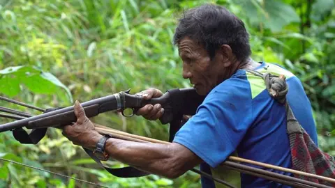 Juan menodongkan pistol dengan kaos biru 