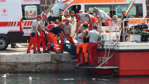 Reuters Layanan darurat membawa kantong jenazah setelah sebuah kapal terbalik Senin pagi di lepas pantai Porticello dekat kota Palermo, Sisilia, Italia, 19 Agustus 2024.