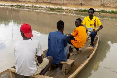 Rektor MBAIRAMADJI MOITA/EPA Masyarakat menggunakan perahu untuk mengarungi jalan yang terendam banjir.