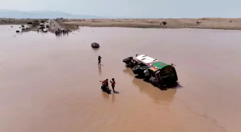     Reuters Orang-orang berdiri di samping kendaraan yang terbalik di tengah banjir di Yaman. 