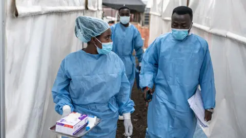 Getty Images Petugas kesehatan yang mengenakan masker dan scrub mengobrol saat mereka berjalan di antara tenda di pusat pengobatan cacar di Dungwe, Kivu Utara, DR Kongo, pada 17 Agustus.