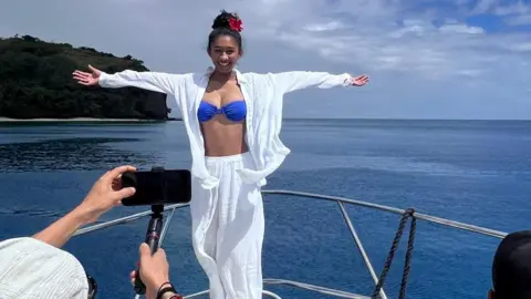 Jennifer Chan Seorang wanita yang mengenakan kemeja putih dan bawahan serta atasan bikini biru berpose di atas perahu di laut sementara seorang pria mengambil foto dengan ponselnya.