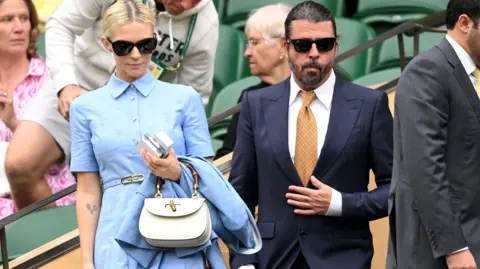 Getty Images Mengenakan gaun berlumuran darah dan memegang tas tangan putih di tangan kirinya, Jordyn Blum berdiri di samping suaminya Dave Grohl saat mereka duduk di kejuaraan tenis Wimbledon. Grohl mengenakan setelan biru tua, kacamata hitam, dan dasi oranye.