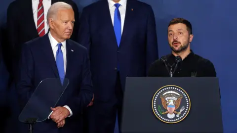 Mengenakan dasi biru dan setelan biru laut EPA, Presiden AS Joe Biden berdiri di samping Presiden Ukraina Volodymyr Zelensky saat ia berbicara di podium dalam pidatonya pada KTT NATO di Washington, Juli 2024.