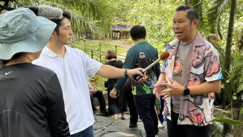 Direktur Kebun Binatang Terbuka Khao Khiev/Kebun Binatang X Narongwit Chodchoi, mengenakan kemeja bermotif kuda nil, memberikan wawancara kepada kru televisi Jepang.