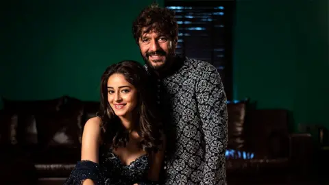 Hindustan Times/Getty Images Ananya Pandey duduk di rumahnya bersama ayahnya Chunky Pandey berdiri di sisinya. Mereka tersenyum dengan pakaian tradisional India yang cerah. Di belakang mereka di ruang tamu ada sofa kulit berwarna coklat dan dinding hijau.