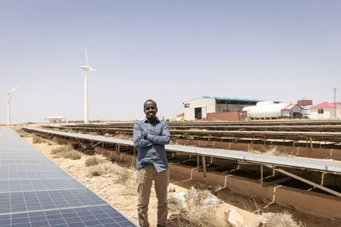 Alyona Synenko/ICRC Salah satu penyedia listrik dan energi terbesar di Somalia, NECSOM adalah produsen bahan bakar fosil hibrida dan energi ramah lingkungan. Di sini seorang pria tersenyum berdiri di tepi panel surya, tangan terlipat di depan dada.