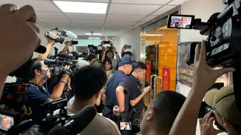 Dua petugas polisi dikelilingi oleh reporter Joy Chiang/BBC saat mereka memasuki kantor Gold Apollo di Taipei. 