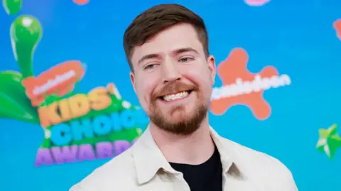 Getty Images Jimmy Donaldson, alias Mr. Beast, berfoto di Nickelodeon Kids' Choice Awards. MrBeast memiliki rambut coklat pendek dan janggut kecil. Dia mengenakan jaket putih di atas T-shirt hitam dan berpose di depan papan iklan biru cerah dengan branding oranye Nickelodeon. 