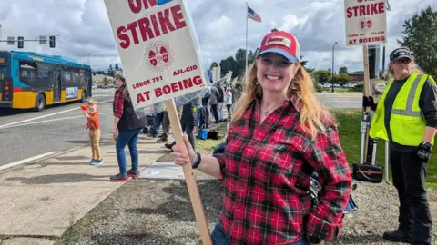 Kerry Foster yang tersenyum, mengenakan kemeja kotak-kotak merah dan topi baseball, memegang tanda mogok di luar Boeing, bendera Amerika terlihat di belakangnya, dan sebuah bus melaju di jalan