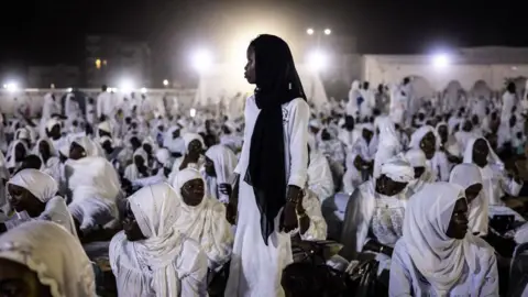 John Wessels/AFP Gambar seorang remaja putri berdiri menghadap cahaya putih terang, di antara perempuan lain berpakaian serba putih, duduk Minggu, 15 September 2024, di Dakar, Senegal.