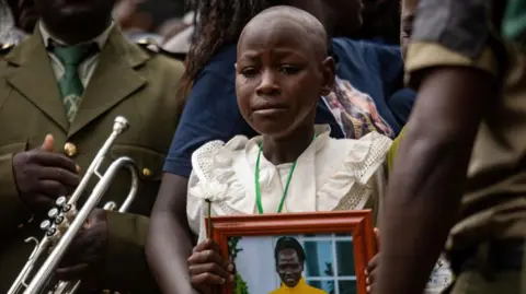 Badru Katumba/AFP Image memperlihatkan putri kecil Rebecca Cheptegei menangis sambil memegang foto ibunya di pemakamannya di distrik Bukwo, Uganda - Sabtu 14 September 2024.