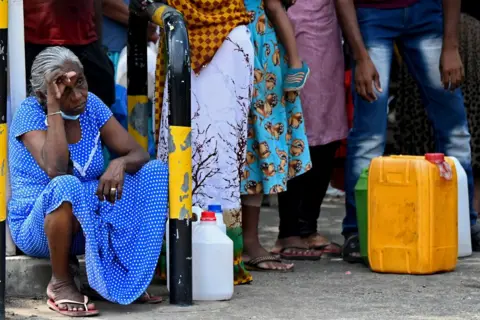 Getty Images Seorang wanita dengan gaun berbintik biru duduk di antrean orang di samping wadah bensin