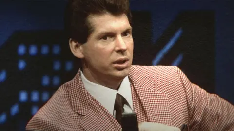 Tn. McMahon/Netflix Vince McMahon muda dari tahun 1980-an atau 1990-an, mengenakan setelan kotak-kotak berwarna merah muda dan putih dengan kemeja putih dan dasi hitam.