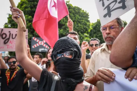 BEN EBRAHIM/GETTY Seorang demonstran perempuan muda, menutupi wajahnya dengan bandana, mengangkat tangannya saat yang lain mengibarkan bendera nasional Tunisia dalam demonstrasi pada 22 September 2024.