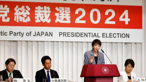 Getty Images Seorang wanita berjas berbicara di mimbar di depan papan bertuliskan 'Pemilihan Presiden 2024' dalam karakter Jepang. Tiga orang berjas, dua pria dan satu wanita, duduk bersamanya.