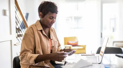 Getty Images Seorang wanita duduk di depan laptop dan melihat sebuah amplop penuh uang kertas