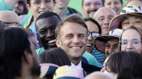 Andre Payne/Pool/AFP Presiden Prancis Emmanuel Macron berpose bersama para sukarelawan saat ia mengunjungi kompetisi panahan selama Olimpiade Paris 2024 di Esplanade des Invalides di Paris pada 2 Agustus 2024.