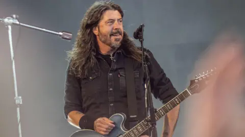 Getty Images Musisi AS Dave Grohl dari band rock Foo Fighters tampil di Orange Stage di Festival Roskilde pada 5 Juli di Roskilde, Denmark. Grohl yang mengenakan kemeja hitam meletakkan tangan kanannya di atas gitarnya saat berpose di depan pertunjukan. Grohl melihat ke arah burung gagak.