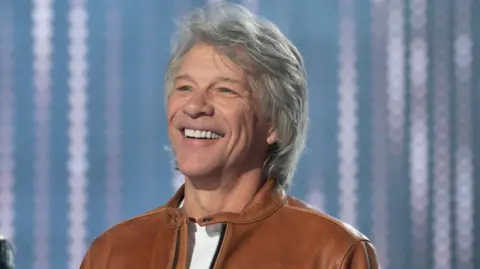 Getty Images Legenda rock AS Jon Bon Jovi tersenyum dengan kaus putih dan jaket kulit cokelat saat tampil di serial American Idol pada Mei 2024.