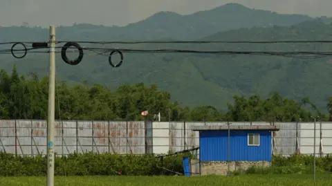 Xiqing Wang/BBC Pagar perbatasan logam yang tinggi membelah ladang antara Tiongkok dan Myanmar di Ruili 