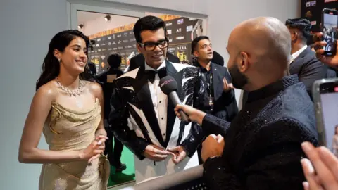 Janhvi Kapoor, Karan Johar dalam tuksedo putih dan hitam dengan gaun emas berkilauan, dasi hitam dan kacamata berbingkai gelap berbicara kepada Haroon Rashid dari Asian Network dengan mikrofon gelap terentang. Haroon mengenakan setelan gelap.