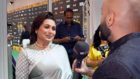 Haroon Rashid, memegang mikrofon gelap, menanyakan pertanyaan kepada Rani Mukerji di karpet hijau IIFA Awards, Rani mengenakan saree abu-abu tradisional berkilauan dan kalung mutiara putih.