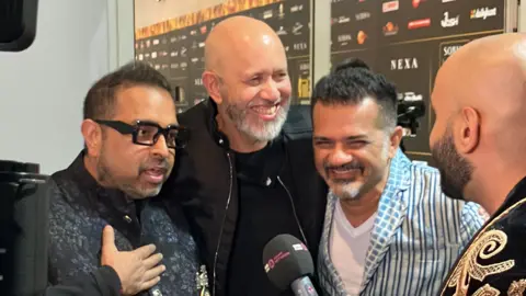 Komposisi Trip Shankar-Ehsaan-Loy tersenyum saat berbicara dengan Haroon Rashid di karpet hijau IIFA Awards. Shankar mengenakan gaun berwarna gelap bermotif bunga dengan lengan di depan dada, Ehsaan dengan pakaian serba hitam, dan Loy dengan kemeja biru bergaris dengan kemeja putih.