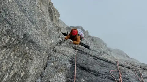 Diambil dari puncak Fay Manners, wanita berbaju oranye, wajahnya tertutup, mendaki permukaan gunung yang jernih, menutupi titik kabut tebal di bawahnya.