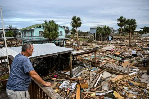 Chandan Khanna/AFP Seorang pria memeriksa kerusakan rumahnya setelah Badai Helen. Gambar tersebut memperlihatkan sampah dari rumah-rumah sejauh mata memandang.