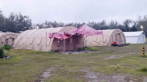 Deretan tenda besar berwarna khaki menampung para migran secara berkelompok, dan beberapa tenda berwarna putih digunakan sebagai ruang komunal darurat.