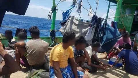 Para migran Tamil, dengan wajah tertutup atau jauh dari kamera, duduk di dek kapal, mencuci tali di sekeliling mereka, saat mereka berlayar melintasi Samudera Hindia.