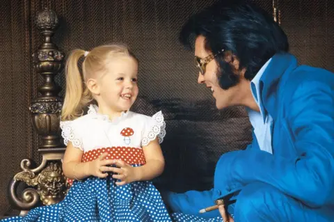 EPE, Inc Lisa Marie, usia 2 tahun, dan Elvis difoto oleh Frank Carroll di rumah keluarga mereka di Hillcrest, LA pada bulan November 1970. Dia menatap lurus ke arahnya dan tersenyum. Dia memiliki rambut pirang dikuncir dan gaun polkadot merah, biru dan putih dengan gambar jamur di atasnya. Elvis mengenakan setelan biru cerah, dia memegang cerutu, dia memiliki cincin besar di jari kelingkingnya, dan dia memakai kacamata hitam. Dia tersenyum lurus padanya.