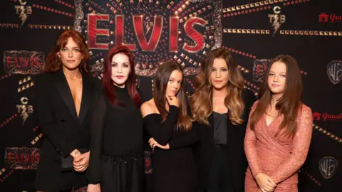 Getty Images untuk Warner Bros. Riley Keough, Priscilla Presley, Harper Vivienne Ann Lockwood, Lisa Marie Presley dan Finley Aaron Love Lockwood pada pemutaran khusus ELVIS pada 11 Juni 2022. Semua orang berpakaian hitam kecuali Finley yang mengenakan Peachy. - Gaun renda merah muda. Elvis berdiri di depan poster iklan film.