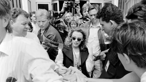 Getty Images Foto hitam putih menunjukkan Kylie Minogue dikerumuni penggemar dan paparazzi di luar studio Capital Radio di London pada tahun 1989