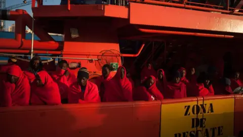 Getty Images Para migran dan pengungsi yang mengenakan selimut untuk menghangatkan tubuh berdiri di atas kapal yang dioperasikan oleh Layanan Penyelamatan Maritim Spanyol