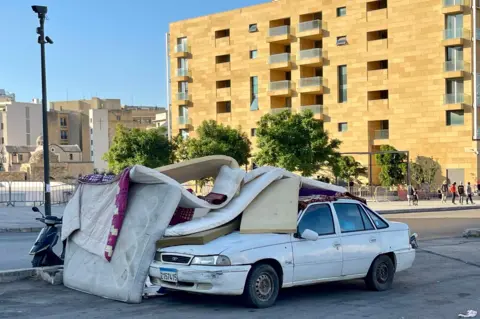 Sebuah mobil dengan kasur di atasnya, di depan sebuah blok apartemen