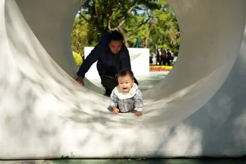 Seorang bayi merangkak melalui terowongan di taman, dengan seorang wanita dewasa di belakangnya