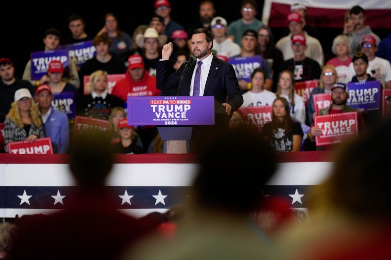 JD Vance berdiri di belakang podium bermerek Trump dan berbicara kepada orang banyak.