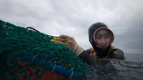 Apple A Henyo, penyelam dari Pulau Jeju Korea Selatan dalam The Last of the Sea Woman, mengenakan sarung tangan pelindung berwarna kuning dan perlengkapan menyelam sambil memegang jaring ikan berwarna hijau