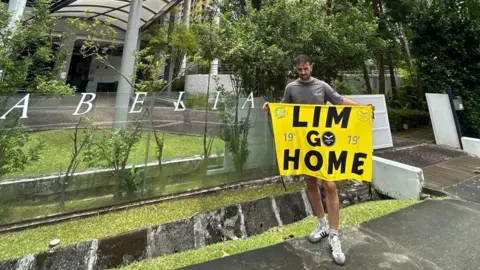 Dani Cuesta/XA Seorang pria di luar kompleks apartemen perumahan memegang spanduk kuning bertuliskan LIM GO HOME